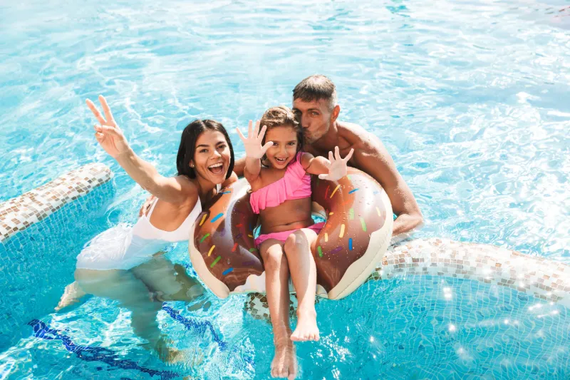 En glad familj bestående av en mamma, pappa och en ung dotter som badar i en solig pool. Dottern sitter i en uppblåsbar badring formad som en munk.
